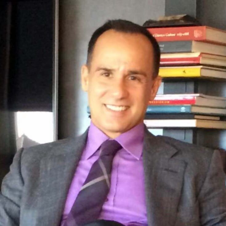 A man in purple shirt and jacket sitting next to books.
